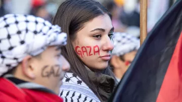 L'image montre des manifestants participant à une manifestation, probablement en lien avec la situation à Gaza. Une jeune femme a le mot "Gaza" peint en rouge sur sa joue, tandis qu'une autre personne à côté d'elle porte un foulard traditionnel palestinien et a également "Gaza" écrit sur son visage, mais en noir. Ils sont entourés d'un public qui semble mobilisé pour une cause. L'atmosphère est sérieuse, reflétant un engagement fort pour la situation en Palestine.