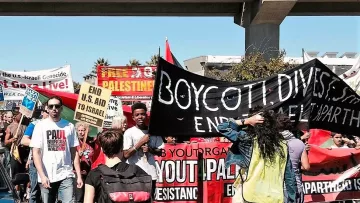 L'image montre une manifestation avec de nombreuses personnes tenant des pancartes et des bannières. Les participants semblent exprimer des opinions politiques concernant Israël et la Palestine. On peut lire des slogans comme "Boycott, Désinvestissez, Sanctionnez" et "Finissez avec Israël". L'ambiance paraît dynamique, avec des gens engagés et un sentiment d'unité pour une cause. La scène se déroule sous un viaduc, ajoutant au cadre urbain de l'événement.