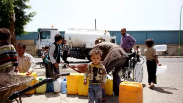 L'image montre une scène animée dans une zone urbaine, où plusieurs personnes sont rassemblées autour d'un camion-citerne. Des adultes et des enfants transportent des récipients pour collecter de l'eau. On remarque un jeune enfant au premier plan, portant une veste colorée, se tenant debout parmi d'autres personnes qui s'affairent. En arrière-plan, on aperçoit un homme en fauteuil roulant qui semble aider. Les couleurs des récipients sont vives, principalement jaunes et bleus, et l'atmosphère illustre un besoin urgent d'approvisionnement en eau.