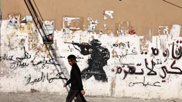 A boy walks past a wall with a soldier graffiti and Arabic text, contrasting street life.