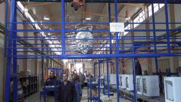 The image shows an industrial setting, likely a manufacturing or assembly facility. There are several workers in the foreground, wearing uniforms, and they seem to be engaged in tasks related to assembling or inspecting products. Above them, there is overhead equipment, possibly a crane or hoist, used for lifting materials. The facility has rows of workstations with what appears to be appliances or machinery on display, indicating a focus on production. The interior is well-lit with a spacious design, featuring high ceilings and a structured layout.