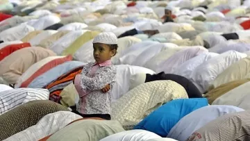 L'image montre un jeune garçon, vêtu d'une chemise à motifs et d'une petite kufi blanche, se tenant debout avec les bras croisés. Il regarde autour de lui, tandis qu'autour de lui, un grand nombre de personnes sont en prière, les corps inclinés et recouverts de vêtements colorés. L'ambiance semble empreinte de sérénité et de dévotion, avec des draps et des vêtements disposés sur le sol, typiques d'un rassemblement de prière.