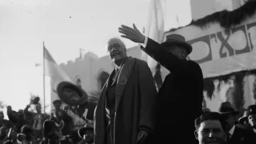 L'image montre des hommes en costume debout sur une estrade, saluant une foule rassemblée. L'un des hommes, portant un manteau, semble s'adresser au public, tandis que l'autre, en chapeau, lève la main. En arrière-plan, on aperçoit des drapeaux et des décorations, indiquant un événement officiel ou une célébration. La foule est composée de personnes applaudissant et prenant part à l'événement. L'atmosphère semble festive et solennelle.