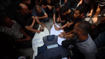 A crowd mourns over a deceased person covered in a white cloth, wearing a press vest.
