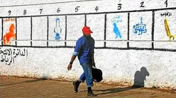 L'image montre un homme marchant le long d'un mur peint en blanc. Sur le mur, il y a une série de numéros de 1 à 8 avec des illustrations ou des symboles correspondant à chaque numéro. L'homme porte une casquette rouge et un vêtement bleu, tenant une mallette. À gauche du mur, une inscription en arabe est visible. L'environnement semble urbain et ensoleillé.