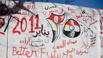 The image features a wall covered in graffiti and artwork. Prominent elements include the Egyptian flag, the date "25 January 2011," and references to the revolution. Phrases in Arabic and English express themes of hope, such as "Better Future" and "إرادة," meaning "will" or "determination." The background suggests a lively and politically charged atmosphere, symbolizing the spirit of change during the Egyptian revolution. Various colors like red, black, and white are used, enhancing the visual impact of the message.