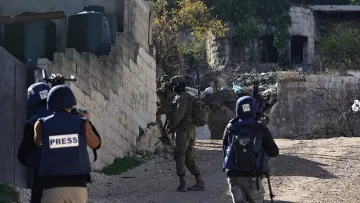 L'image montre une scène de conflit avec des soldats armés et des journalistes. Il y a des soldats en uniforme, portant des casques et des équipements de protection, qui se déplacent dans un environnement urbain, probablement dans une zone de conflit. Des journalistes sont également présents, identifiables grâce à leurs gilets marqués du mot "PRESS". En arrière-plan, on peut apercevoir des structures en ruine et des éléments de débris, ce qui suggère un contexte de tensions ou de violence.