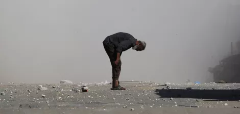 مردی مسن با کمر خمیده در مکانی خاکی و غبارآلود ایستاده است. اطرافش صحنه‌ای ویران وجود دارد.