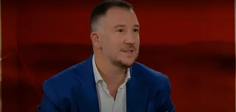 L'immagine mostra un uomo con capelli corti e una barba leggera, che indossa una giacca blu e una camicia bianca. Si trova di fronte a uno sfondo rosso. L'uomo appare in un contesto di colloquio o discussione, con un'espressione attenta e coinvolta.