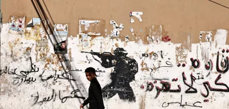 A boy walks past a wall with a soldier graffiti and Arabic text, contrasting street life.