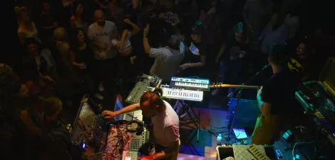 A lively crowd enjoys live music from performers with electronic instruments on stage.