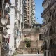 L'image montre une vue urbaine d'une rue avec des bâtiments en pierre et des murs en béton. On observe un mélange d'architecture ancienne et moderne, avec des balcons ornés de fer forgé. De nombreux paraboles satellites sont installées sur les façades des bâtiments, tandis que des linges pendus à l'extérieur ajoutent une touche de vie quotidienne. Les escaliers menant à d'autres niveaux sont visibles, et le ciel est clair et ensoleillé, donnant une atmosphère vive à la scène.