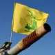 L'image montre un canon pointant vers le ciel, avec un drapeau jaune flottant au-dessus. Sur le drapeau, il y a des inscriptions en arabe ainsi qu'un logo symbolique. Le fond est bleu, suggérant une journée ensoleillée. Le canon présente des signes d'usure, avec une surface rouillée.