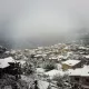 L'image montre un paysage hivernal où une petite ville est recouverte de neige. Les toits des maisons, en grande partie en bois, sont également blancs, créant une atmosphère paisible et froide. On peut apercevoir des arbres chargés de neige et des collines en arrière-plan, le tout enveloppé par une brume légère, ajoutant à la sérénité de la scène. La vue donne une impression de calme et d'isolement, typique des régions montagneuses en hiver.