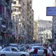 L'image montre une rue animée dans une ville, probablement une métropole. On peut voir de nombreux bâtiments de plusieurs étages qui bordent la route. Les voitures sont garées et circulent, créant un certain embouteillage. Une foule de personnes se déplace le long des trottoirs. Des panneaux de signalisation et des enseignes en arabe sont visibles, indiquant que cette scène se déroule dans un pays arabophone. L'atmosphère semble dynamique et vivante, typique d'une grande ville.