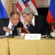 L'image montre deux hommes assis à une table, engageant une conversation. L'un est identifié comme étant le ministre des Affaires étrangères de la Russie, Sergueï Lavrov, et l'autre comme John Kerry, l'ancien secrétaire d'État américain. Ils semblent discuter de manière confidentielle, entourés de décorations représentant les drapeaux de la Russie et des États-Unis. Sur la table, il y a des documents et une tasse. L'atmosphère semble sérieuse et diplomatique.