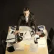 L'image montre un homme debout sur une table en bois, entouré de divers documents et d'objets. Il a une expression sérieuse et semble concentré. Sur la table, on peut voir des cartes, des notes, des dossiers et un lampadaire qui éclaire la scène. L'environnement autour de lui est plutôt sobre, avec des chaises noires et un sol sombre. L'ensemble donne une impression de travail acharné et de recherche.