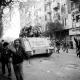 L'image montre une scène de manifestation urbaine, avec des gens dans une rue animée. Au premier plan, plusieurs manifestants marchent, certains levant les bras en l'air. À l'arrière-plan, un tank est visible, portant des inscriptions. La scène est en noir et blanc, ce qui intensifie l'atmosphère dramatique et chargée d'émotion. Les bâtiments environnants et les arbres ajoutent à l'environnement urbain. L'ensemble dégage un sentiment de tension et de mouvement.