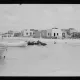 L'image montre une scène maritime où plusieurs bateaux, probablement des barques, sont amarrés au bord d'une eau peu profonde. En arrière-plan, on peut apercevoir des bâtiments en ruines, suggérant une ancienne architecture. À proximité, un homme semble se trouver dans l'eau, tandis qu'un autre est à cheval ou monté sur un animal aquatique. Le ciel est nuageux, apportant une lumière diffuse à l'ensemble de la scène. L'atmosphère semble tranquille et un peu nostalgique, évoquant un passé révolu.