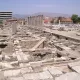 L'image montre un site archéologique avec des ruines et des structures anciennes. On aperçoit des colonnes dressées, probablement d'un bâtiment antique, ainsi que des fondations de murs en pierre. En arrière-plan, on peut distinguer des bâtiments modernes, contrastant avec les vestiges historiques. Le ciel est clair et le paysage est légèrement vallonné au loin. L'endroit semble être une zone de fouilles, avec des éléments qui témoignent d'une ancienne civilisation.