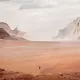 L'image montre un vaste paysage désertique. On peut voir des montagnes rocheuses au loin, entourées de dunes de sable. Le ciel est nuageux, ce qui crée une atmosphère légèrement brumeuse. Au centre, une seule silhouette humaine, probablement un cavalier, se déplace à travers ce paysage aride. La scène transmet une sensation d'immensité et de solitude.