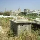 L'image montre un cimetière abandonné ou négligé, où des tombes en béton sont entourées d'herbes hautes et de végétation. On peut apercevoir, au fond, des bâtiments de plusieurs étages et des structures urbaines, ce qui indique une proximité avec une zone habitée. L'atmosphère est calme, mais on ressent un contraste entre l'état de mise à l'écart des tombes et l'activité de la ville environnante.