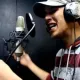 L'image montre un homme en train de chanter ou de rapper dans un studio d'enregistrement. Il porte un chapeau et des écouteurs. Devant lui, un microphone est connecté et il tient des feuilles de papier, probablement des paroles ou un texte. L'expression de son visage indique qu'il est très impliqué dans sa performance. L'arrière-plan semble sombre, ce qui est typique des environnements d'enregistrement.