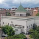 L'image montre un bâtiment architectural distinctif avec une façade blanche et un toit en tuiles vertes. Le design présente des éléments inspirés de l'architecture islamique, comme des dômes et des arches. En arrière-plan, on aperçoit des immeubles de style haussmannien, typiques de nombreuses villes françaises, avec des balcons et des toits en attique. Le ciel est nuageux, ce qui ajoute une atmosphère particulière à la scène. Des arbres et de la verdure entourent le bâtiment, contribuant à l'esthétique générale.