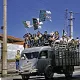 L'image montre un camion dont le plateau est rempli de personnes célébrant avec des drapeaux algériens. Les gens semblent joyeux et enthousiastes, tenant les drapeaux en l'air. En arrière-plan, on aperçoit des bâtiments et des pylônes électriques. La scène évoque une ambiance festive et patriotique. Les couleurs sont vives, et l'ensemble transmet un sentiment d'unité et de célébration.