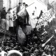 L'image montre une grande foule rassemblée, avec des individus brandissant des drapeaux et des bannières. L'atmosphère semble tendue et chargeée d'émotion, probablement liée à un événement historique. Des personnes se tiennent sur un bâtiment, tandis que d'autres sont regroupées en bas, certaines portant des uniformes, ce qui pourrait indiquer une présence policière ou militaire. Le message sur les bannières évoque des thèmes d'indépendance, suggérant un contexte de luttes sociales ou politiques.