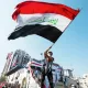 L'image montre une personne agitant un drapeau irakien. Le drapeau, aux couleurs rouge, blanc, noir et vert, est bien visible. La scène se déroule probablement lors d'une manifestation ou d'un événement public, avec des bâtiments en arrière-plan décorés de drapeaux. L'atmosphère semble dynamique, reflétant un sentiment de patriotisme et de rassemblement.