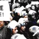 L'image montre une scène de manifestation où un homme tient une grande pancarte avec l'inscription "PEUPLE DÉGAGE !" Au premier plan, cet homme semble déterminé et engagé, tandis qu'en arrière-plan, un groupe de policiers en uniforme avec des casques se tient à proximité, suggérant une tension entre les manifestants et les forces de l'ordre. L'atmosphère semble chargée d'émotion et de revendication.