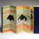 L'image montre un livre artistique aux pages colorées, présentant des illustrations de montagnes. Les pages sont pliées en accordéon et comportent des inscriptions en arabe. La palette de couleurs est variée, avec des nuances de jaune, bleu et orange, ajoutant une dimension vibrante à l'ensemble. Le livre est partiellement ouvert, révélant sa structure unique et sa conception créative. Une couverture noire est visible à droite, contrastant avec les couleurs vives des pages intérieures.