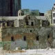 L'image montre des ruines d'un bâtiment ancien, probablement une église, dont la structure est en pierre et partiellement en décomposition. La façade est endommagée et recouverte de végétation, tandis qu'en arrière-plan, on peut voir des immeubles modernes en construction ou en rénovation. Le contraste entre l'ancien et le moderne donne une atmosphère de dégradation et de changement architectural. La zone semble être en chantier, avec des barrières et des échafaudages visibles.