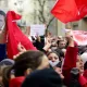 L'image montre une foule de manifestants rassemblés, brandissant des drapeaux rouges et des pancartes. Certains visages sont masqués, tandis que d'autres expriment des émotions intenses, comme la détermination ou la colère. On aperçoit aussi un portrait d'une figure emblématique en arrière-plan. Les slogans sur les pancartes semblent appeler au changement et à la liberté, indiquant un contexte de protestation politique. L'atmosphère est chargée d'énergie collective.