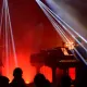 L'image montre une scène de concert avec deux musiciens. À gauche, un batteur joue de la batterie, tandis qu'à droite, un pianiste est assis devant un clavier. L'éclairage est dramatique, avec des faisceaux lumineux qui coupent à travers la fumée ambiante, créant une atmosphère dynamique. En arrière-plan, on devine une audience silhouette, profitant du spectacle. Les couleurs rouges et jaunes ajoutent à l'énergie de la performance.