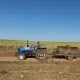 L'image montre un homme conduisant un tracteur bleu sur une route de terre. Il tire une remorque avec un grand réservoir métallique. À l'arrière-plan, on peut voir des champs verts sous un ciel dégagé. La scène évoque une ambiance rurale et agricole.