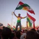 L'image montre une grande foule célébrant, probablement lors d'un événement culturel ou politique. Au premier plan, un homme se tient debout sur une autre personne, tenant des drapeaux kurdes. Il porte un maillot aux couleurs du drapeau kurde, et son expression semble joyeuse et enthousiaste. En arrière-plan, on peut voir de nombreuses autres personnes agitant également des drapeaux, créant une atmosphère festive et énergétique. Le ciel est clair, ce qui ajoute à l'ambiance dégagée de la scène.