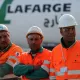 L'image montre trois ouvriers portant des vêtements de sécurité orange et des casques blancs. Ils se tiennent debout avec les bras croisés, affichant une attitude sérieuse. En arrière-plan, on peut voir un camion ou un véhicule avec le logo de Lafarge, une entreprise connue dans le secteur des matériaux de construction. L'environnement semble être un chantier ou une zone industrielle.