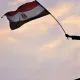 L'image montre une silhouette d'une personne debout sur une plateforme, levant un drapeau. Le drapeau est égyptien, avec des couleurs rouge, blanc, noir et le symbole du pays. L'arrière-plan présente un ciel avec des nuages aux teintes douces, créant une ambiance de détermination et de célébration. Cette scène évoque des sentiments de patriotisme et de protestation ou de victoire.