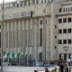 L'image montre un bâtiment majestueux avec une architecture moderne. La façade est claire, avec de grandes fenêtres et des lignes géométriques. On peut également voir des drapeaux algériens flottant devant le bâtiment. Il y a des lampadaires et une ambiance urbaine. Ce contraste entre le style architectural et le décor environnant suggère un lieu important, probablement lié aux institutions gouvernementales.