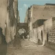 L'image représente une ruelle étroite et pavée, probablement dans une ville historique. Les murs des bâtiments sont en pierre, avec des textures vieilles et usées, suggérant un certain âge. On peut apercevoir des silhouettes de personnes marchant, certaines vêtues de longues robes traditionnelles. À l'arrière-plan, un arc architectural est visible, ajoutant une touche d'authenticité à l'environnement. Le ciel est dégagé, apportant une lumière douce à la scène. L'atmosphère semble paisible et évoque un lieu chargé d'histoire.