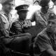 L'image montre un groupe de militaires assis dans un véhicule. Il y a plusieurs hommes en uniforme, portant des chapeaux distinctifs. Certains sont assis à l'avant, tandis que d'autres semblent être à l'arrière. L'atmosphère est solennelle, et il pourrait s'agir d'un moment lié à une opération ou une mission militaire. Le fond est flou, mais on aperçoit d'autres véhicules et un environnement urbain.