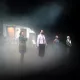 L'image présente une scène de théâtre avec quatre personnages en position debout sur scène. Ils se trouvent dans une atmosphère brumeuse, ce qui ajoute une ambiance mystérieuse. En arrière-plan, on peut apercevoir une petite maison ou un décor qui semble inhabitée. Les personnages sont vêtus de manières diverses, ce qui suggère des rôles ou des traits de caractère différents. La lumière semble tamisée, renforçant l'effet dramatique de la scène.