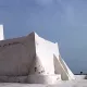 L'image montre un bâtiment de couleur blanche, typiquement de style architectural méditerranéen. Il semble être une petite structure, possédant une forme irrégulière avec des murs épais et angulaires. À son sommet, il y a une cloche ou un petit clocher. L'arrière-plan est plutôt clair, avec un ciel bleu ou nuageux qui met en valeur la luminosité de l'édifice. Ce type de construction pourrait évoquer des lieux historiques ou religieux, typiques de certaines régions côtières.