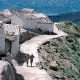 L'image montre un paysage rural avec un sentier serpentant le long d'une colline. De chaque côté du chemin, on peut voir des maisons blanches avec des toits en tuiles. Deux personnes marchent sur le sentier, profitant de la vue. En arrière-plan, des montagnes verdoyantes s'élèvent, créant une atmosphère paisible et naturelle. Le ciel est clair et contribue à l'ambiance sereine de la scène.