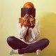 L'image montre une personne assise en tailleur, tenant un livre près de son visage, cachant ainsi son identité. Elle porte un hijab orange et une tenue simple, avec des jeans. L'arrière-plan est d'une couleur claire, créant une ambiance douce et calme. Les bras sont repliés et des bracelets colorés ornent ses poignets. Cette image évoque un moment de réflexion ou de lecture.