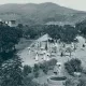 L'image semble représenter une scène rurale d'une époque passée. On peut voir un chemin central bordé d'arbres et de bâtiments à l'arrière-plan. Plusieurs personnes, vêtues de manière traditionnelle, se déplacent, certaines portant des parapluies. Des animaux comme des bœufs sont également présents, ajoutant à l'atmosphère agricole de la scène. Au premier plan, il y a un puits, probablement utilisé par les villageois. Les montagnes en arrière-plan complètent le paysage verdoyant. Cette image évoque une vie paisible et communautaire en harmonie avec la nature.