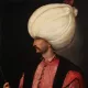 L'image représente un homme en profil, portant un turban blanc imposant. Il est habillé d'une tunique rose ornée de boutons et d'un veston noir, ce qui lui donne un aspect richement décoré. Son visage est finement dessiné, mettant en valeur sa moustache. Il tient un bâton dans une main, ce qui suggère une position de pouvoir ou d'autorité. L'arrière-plan est sombre, ce qui fait ressortir les détails de sa tenue et de son expression.