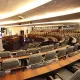 L'image montre une salle de réunion ou un hémicycle, probablement utilisée pour des sessions parlementaires ou des assemblées. On peut voir des rangées de sièges confortables disposés en arc de cercle, avec des tables en bois devant chaque siège. Des écrans et des équipements audiovisuels sont visibles, indiquant que la salle est équipée pour des présentations ou des débats. Les murs sont décorés de manière sobre et moderne, créant une ambiance institutionnelle.